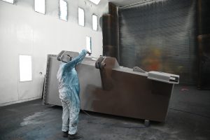 A Marine Travelift employee in an industrial paint suit sprays a large structural steel component in a large paint booth.