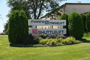 Signs reading Marine Travelift and Shuttlelift outside the factory building in Sturgeon Bay, WI, framed by grass and trees.
