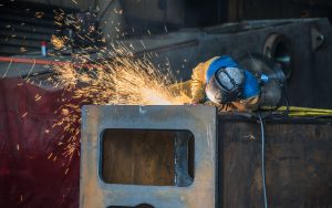 Sparks fly as an ExacTech employee grinds the edge of a large piece of structural steel in a fabrication shop.