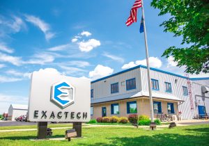 Exactech sign outside the factory building in Sturgeon Bay, WI with green grass, a blue sky, and a waving American Flag