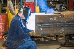 An ExacTech employee in a welding mask crouches next to a large structural steel component to weld
