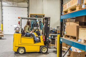 A female employee drives a yellow forklift inside a warehouse space full of shelves with boxes and crates